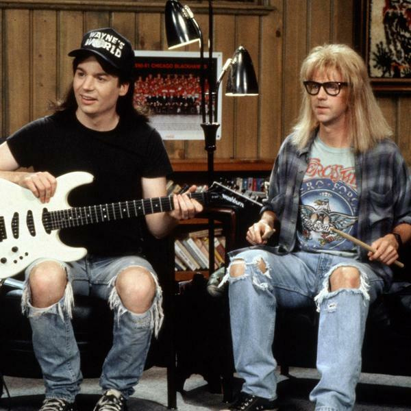 Wayne and Garth sit on a sofa presenting their TV show. Wayne is holding a white guitar, and Garth is holding some drumsticks.